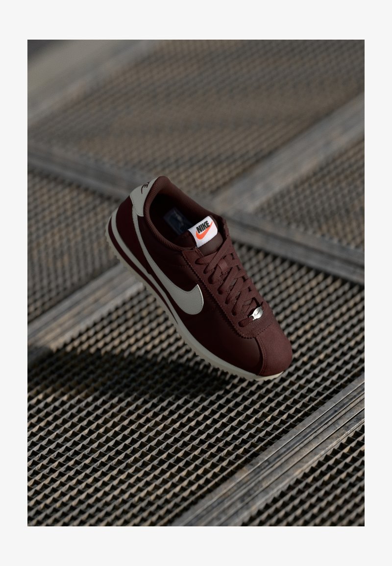 Nike sportbačiai su bordo spalvos viršumi, kreminiu swoosh'u, raištelių užsegimu ir baltais padais, padėti ant tekstūruoto paviršiaus.