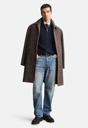 Brown plaid overcoat, navy zip-up sweater, patterned tie, blue denim jeans, black footwear. Focus on textures and layered look.