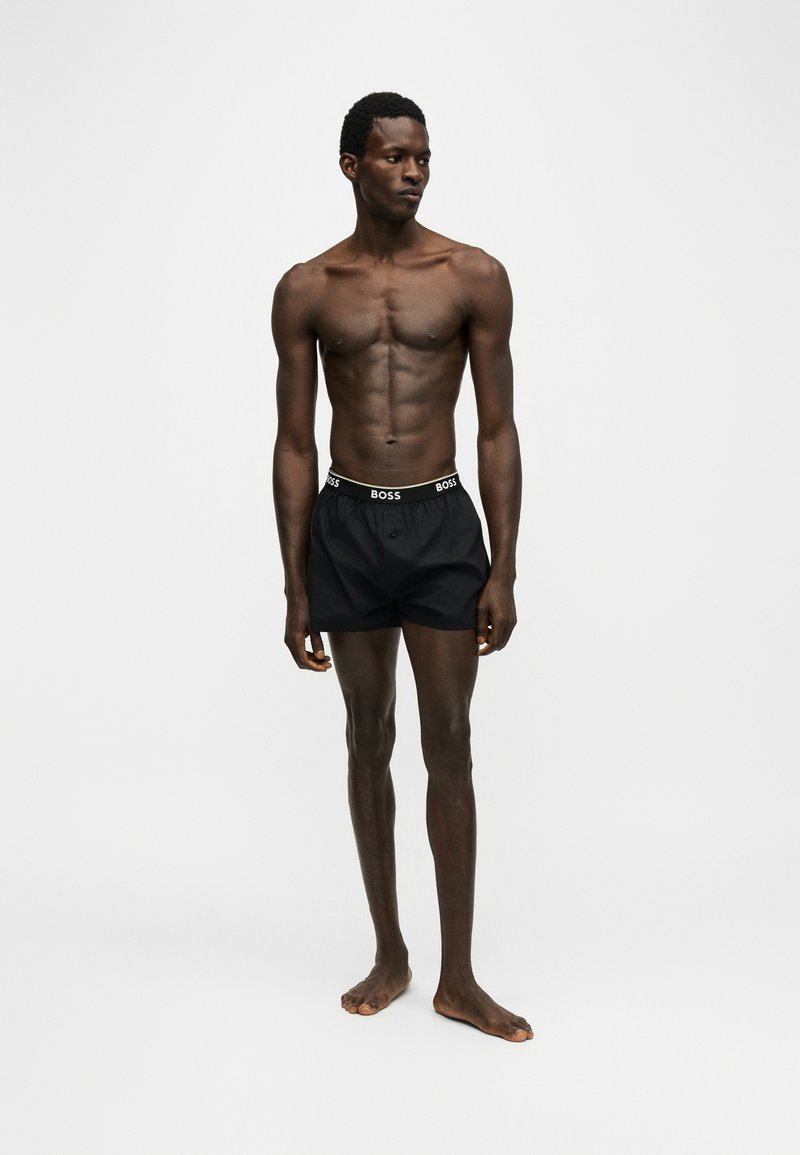Homme mince debout, pieds nus, portant un boxer noir BOSS sur un fond uni gris clair.
