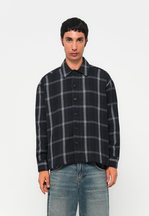 Junger Mann in einem schwarz-weißen karierten Hemd mit Knöpfen und hellblauen Jeans vor einem schlichten weißen Hintergrund.