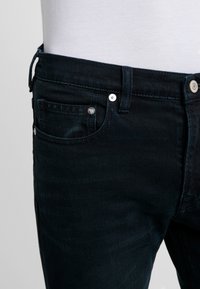 Dark denim jeans with a slim fit, featuring a front pocket, metal button, and contrasting stitching. Material has a slightly textured appearance.