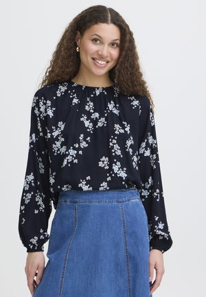 Eine junge Frau mit lockigem Haar trägt eine schwarze, langärmelige Bluse mit Blumenmuster, die in einen hoch taillierten blauen Jeansrock gesteckt ist, und lächelt in die Kamera.