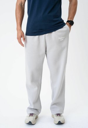 Mand iført lysegrå joggingbukser med teksten "Toujours Actif", navyblå t-shirt, beige sneakers og sort armbåndsur med hånd i lommen.
