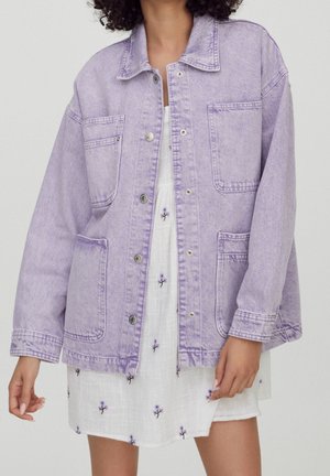 Woman wearing a light purple denim jacket with large front pockets over a white dress embroidered with small purple flowers.