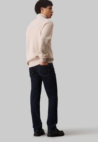 Pull enroulé beige clair à col montant, associé à un jean en denim foncé. Le jean présente des poches arrières et des détails de couture discrets.