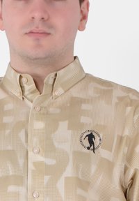 Camicia button-up beige con un motivo testurizzato caratterizzato da grandi elementi grafici e un logo sul lato sinistro del petto. Tessuto morbido, colletto classico.
