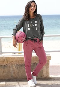 Graues Langarmshirt mit weißem und goldenem Text, kombiniert mit pinken, schmal geschnittenen Jogginghosen und weißen Turnschuhen. Hält einen rosa Basketball.