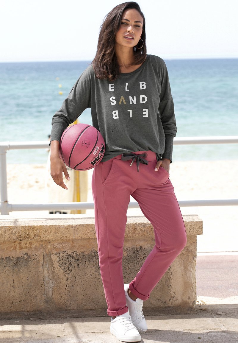Graues Langarmshirt mit weißem und goldenem Text, kombiniert mit pinken, schmal geschnittenen Jogginghosen und weißen Turnschuhen. Hält einen rosa Basketball.