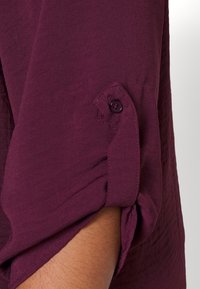 Chemise bordeaux avec les manches retroussées, dotée d'un bouton de fermeture, d'une texture douce et d'un tissu légèrement froissé, mettant en valeur un design décontracté.