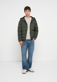 Giacca a vento in nylon verde scuro con cappuccio, indossata sopra un maglione grigio chiaro, abbinata a jeans blu e sneaker bianche.
