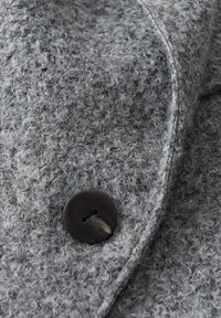 Tissu en laine grise texturé avec des fibres bouclées denses et un gros bouton foncé à deux trous cousu près d'une couture.