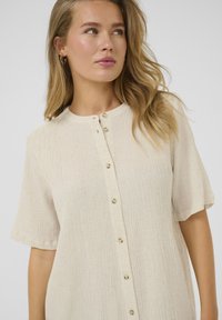 Chemise à bouton légère couleur beige clair avec des manches courtes, tissu texturé, col rond et petits boutons de couleur claire sur le devant.