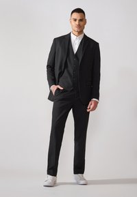 Schwarzer Anzug mit einer taillierten Jacke und passender Hose, über einem weißen Hemd getragen. Weiße Sneakers runden den Look ab und betonen einen modernen Stil.