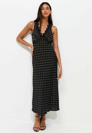 Mujer con vestido maxi negro sin mangas con lunares blancos, borde de encaje, de pie con una mano en la cadera y llevando tacones negros.