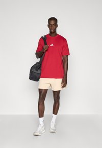 Sarkana īsroku T-krekls ar baltu logotipu, bēšas šorti, melna soma, balti sporta apavi un baltas zeķes. Gluda auduma, brīva piegrieztne, ikdienišķs stils.