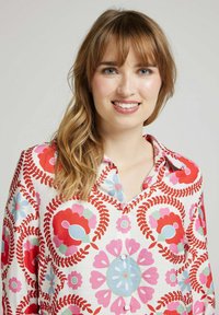 Chemise à motif floral en tissu léger, avec des fleurs rouges et bleues agrémentées de touches vertes. Le col et les détails des boutons sont visibles.