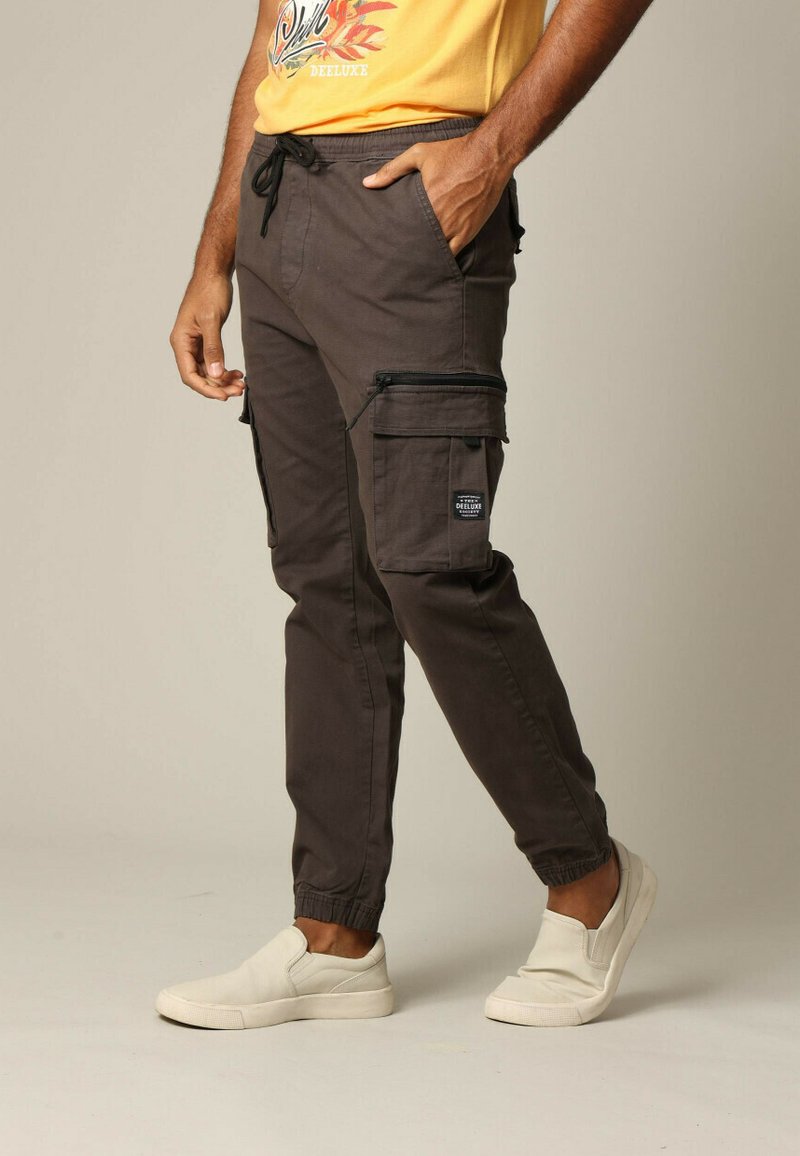 Donkerbruine cargobroek met elastische boorden en een trekkoord in de taille, voorzien van zijritszakken en een patchlogo-detail.