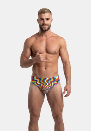 Muskeløs mand med kort blondt hår iført badebukser med regnbuemønster, der laver shaka-håndtegn og smiler mod en hvid baggrund.