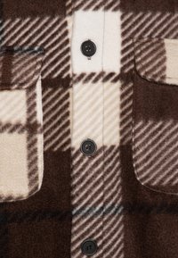 Camisa de franela a cuadros en tonos marrón y beige con botones negros, dos bolsillos en el pecho y un tejido suave y texturizado con un diseño a cuadros.