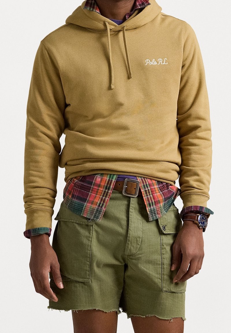 Personne portant un sweat à capuche beige par-dessus une chemise à carreaux rouge et verte, un short cargo vert olive et une montre en cuir marron au poignet gauche.