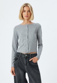 Grauer Langarm-Cardigan mit rundem Ausschnitt, durchgehender Knopfleiste und cropped Design, kombiniert mit hochtaillierten dunklen Jeans.