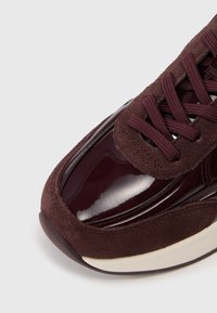 Primo piano di una sneaker bordeaux scuro con punta lucida, sovrapposizioni in camoscio, lacci abbinati e suola bianca su uno sfondo bianco.