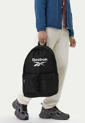 Osoba w białych spodniach i niebieskiej kurtce trzyma czarny plecak Reebok za górną rączkę, nosząc szaro-czerwone trampki.