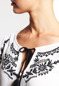 Blouse blanche avec des motifs floraux brodés noirs, un col rond avec un lien, et un tissu léger et texturé.