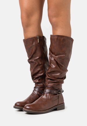 Bottes en cuir marron mi-mollet à enfiler avec fermetures éclair latérales et sangles décoratives portées sur des jambes nues sur fond blanc.