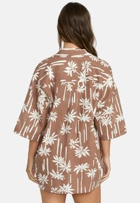 Camicia in lino marrone con stampa di foglie di palma bianche, vestibilità rilassata, maniche corte e dettaglio sulla parte posteriore. Ideale per un look casual.