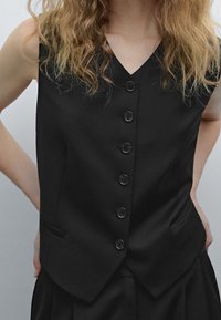 Persona con capelli biondi mossi che indossa un gilet nero senza maniche con bottoni e pantaloni neri, con le mani sui fianchi contro uno sfondo grigio.