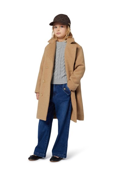 Manteau en laine beige avec des boutons dorés à double boutonnage, pull en maille torsadée gris, jeans large bleu, et casquette à motifs marron. Chaussures noires.