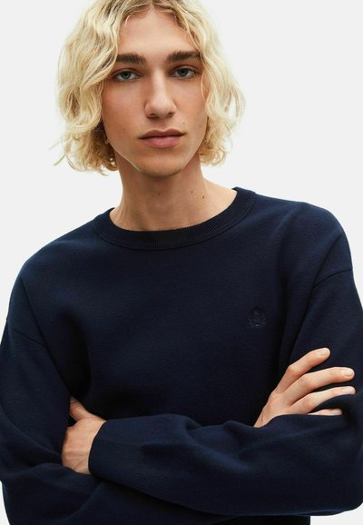 Donkerblauwe gebreide sweater met een geribde ronde hals, een reliëflogo op de borst en een losse pasvorm. De armen van het model zijn gekruist.