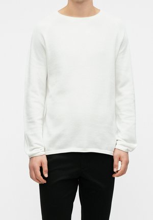 Personne portant un pull texturé blanc à manches longues et un pantalon noir, debout devant un fond blanc uni.