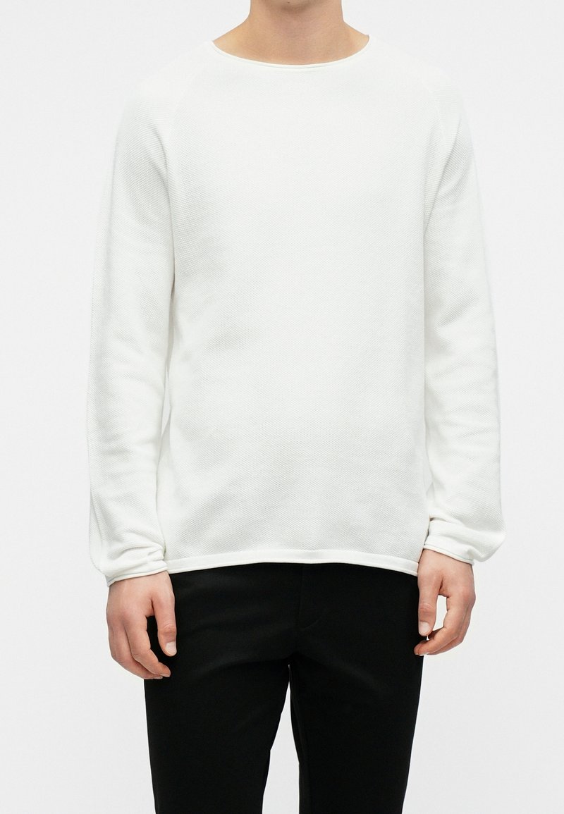 Personne portant un pull texturé blanc à manches longues et un pantalon noir, debout devant un fond blanc uni.