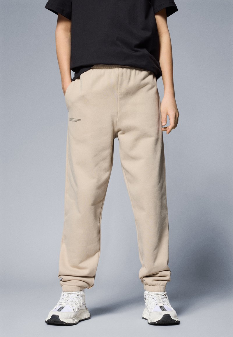 Pantalones de chanda beige con un ajuste relajado, cinturilla elástica y puños. Presentan un detalle de texto minimalista y se combinan con zapatillas deportivas blancas.