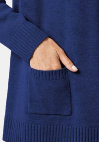 Pull navy avec des poignets et un ourlet côtelés. Comprend une poche frontale avec une bordure texturée. Le matériau semble doux et tricoté.