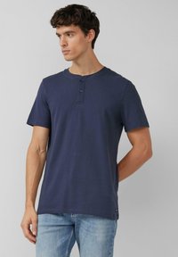 Marineblauw kortermelig henley-shirt van katoen, met een ronde hals, een knoopsluiting met twee knopen en een rechte zoom. Lichtblauwe jeans gedragen.