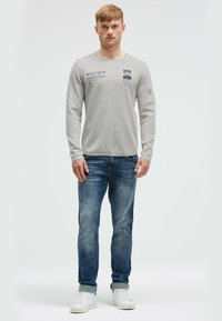 Hellgraues, langärmliges T-Shirt aus strukturiertem Stoff, mit gedruckten Grafiken und Text. Getragen mit blauen Jeans und weißen Turnschuhen.
