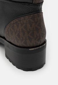 Gros plan sur le talon d'une botte noire et marron en cuir texturé avec une petite plaque métallique gravée Michael Kors.