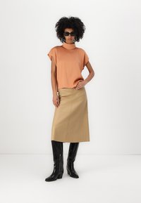 Oransje kortermet topp, beige skinnskjørt, svarte knelange støvler og mørke solbriller. Glatt tekstur og strukturert passform synlig.