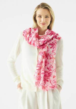 Vrouw draagt een witte cardigan en broek met een roze bloemen sjaal om haar nek, handen in de zakken, tegen een witte achtergrond.