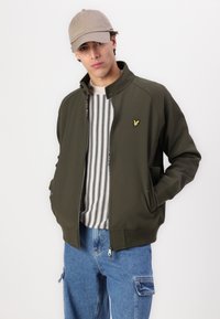 Giacca zip-up verde oliva con colletto a costine, tasche laterali e patch del logo giallo. Indossata sopra un maglione a righe e pantaloni in denim blu.