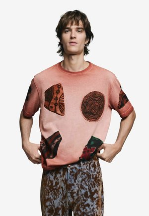 Camiseta de algodón rosa con patrones circulares y abstractos negros. La modelo la lleva con pantalones vaqueros marrones y azules con estampado.