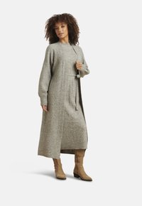 Donna con capelli ricci che indossa un lungo vestito grigio a maglia con motivo a treccia e un cardigan abbinato, abbinati a stivali in suede color cuoio fino a metà polpaccio.