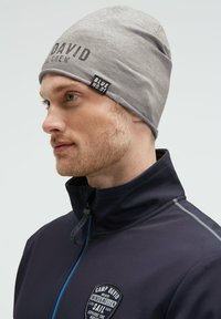 Man with light beard wearing gray beanie labeled "BLUE NO.01" and navy zip-up jacket with Camp David logo patch.