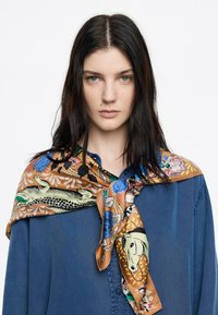 Pañuelo de seda con intrincados patrones de animales y flores en dorado, verde y azul, anudado alrededor de una camisa de mezclilla.