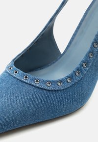 Μπλε ψηλοτάκουνο παπούτσι από τζιν με μυτερή μύτη και ασημένια περσίδα κατά μήκος της άκρης, σε λευκό φόντο.