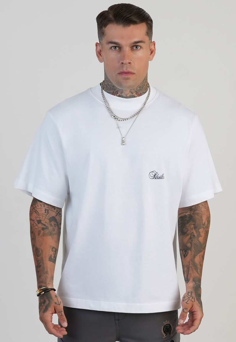 Man with tattoos wearing white oversized T-shirt with small black logo, silver chains, and dark pants against plain background.