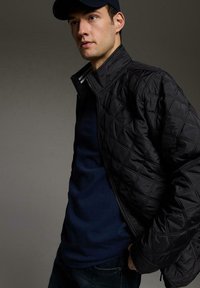 Czarna pikowana kurtka z wysokim kołnierzem, zapinana na zamek, z teksturowanym wzorem, noszona na granatowej koszuli i niebieskich dżinsach. Ciemna czapka dopełnia wygląd.